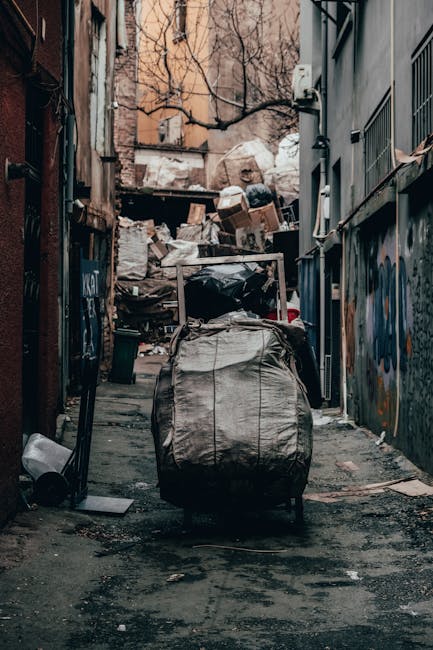 A roadside pile of mixed household waste and refuse, including black plastic rubbish bags, an old beige upholstered armchair with visible wear and tear, a large yellow plastic container, and scattered