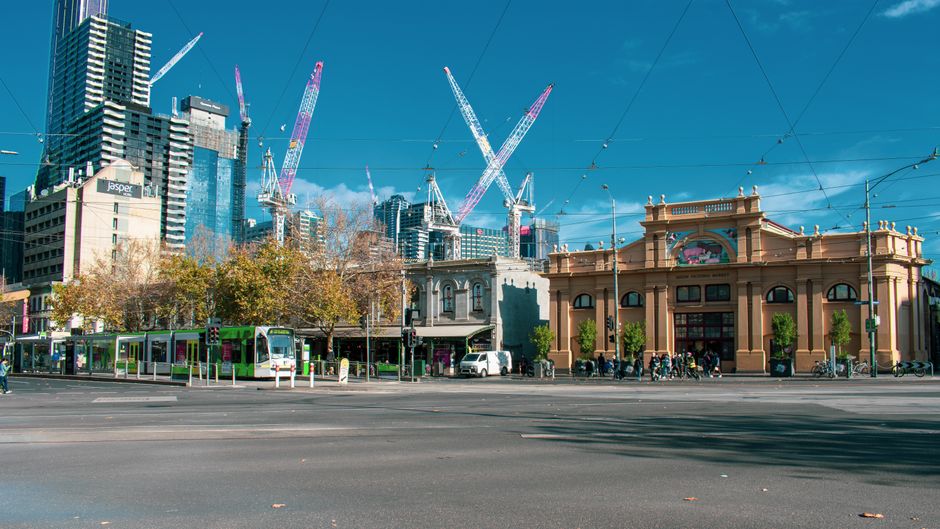 The image depicts a city street scene with a mix of historic and modern buildings, featuring intricate brick facades and large, arched windows. In the foreground, a light blue waste collection truck i